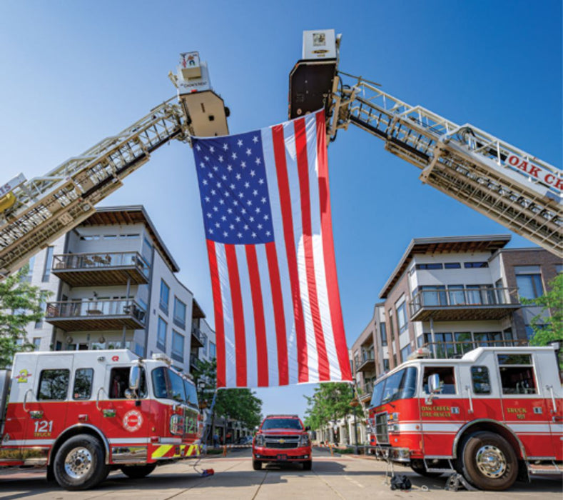 A large American flag hangs between the extended ladders of two fire trucks parked on a city street with apartment buildings in the background.