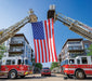 A large American flag hangs between the extended ladders of two fire trucks parked on a city street with apartment buildings in the background.